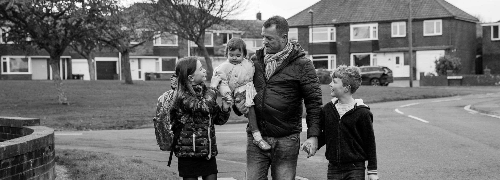 A father holding the hands of two of his school-age children while holding a baby with a typical set off cottage flats int he background.